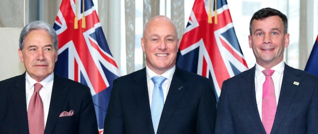 A cropped version of a photo from the press conference to announce the Coalition Agreement published by the spinoff featuring a background of greige vertical blinds and two hanging NZ lags. In the foreground framed head and shoulders view, standing in a row, from left, Winston Peters is dapper in an impeccable dark suit, set off by a very nicely detailed crisp white shirt, a beautifully proportioned and perfectly knotted luxuriant red tie and rakishly arranged, matching pocket square. He is looking somewhat grimly sideways at Luxon beside him. Luxon is all sharp angles and shiny skin, we can see lots of teeth but his smile looks strained, his shiny pale blue tie is absolutely the wrong colour for him, it washes him out, one could be forgiven for imagining perhaps he's even just a tad nauseous knowing people will find out what an epic sock puppet he is. A very ordinary suit, the shoulders don't seem cut quite right for him, its all shiny angles, the tie, collar and neckline gives a blocky vertical chyron look. Shiny suit, shiny smile. Thank god we can't see the pants. To his right is David Seymour. I think he's smiling but it's hard to tell. His suit is slightly lighter than the other two, (all in varying shades of blue), and an edgier, better fitting cut. Unfortunately the jacket is let down by the shirt, which looks unironed and the ugly squat knot on the borderline ugly pink tie.